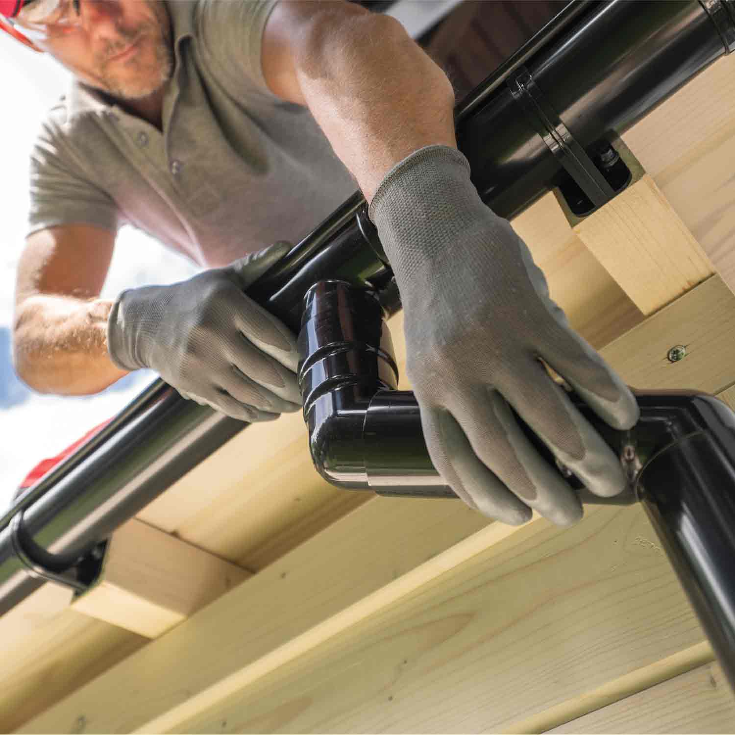 Gloved worker installs black rain gutter on wooden roof.