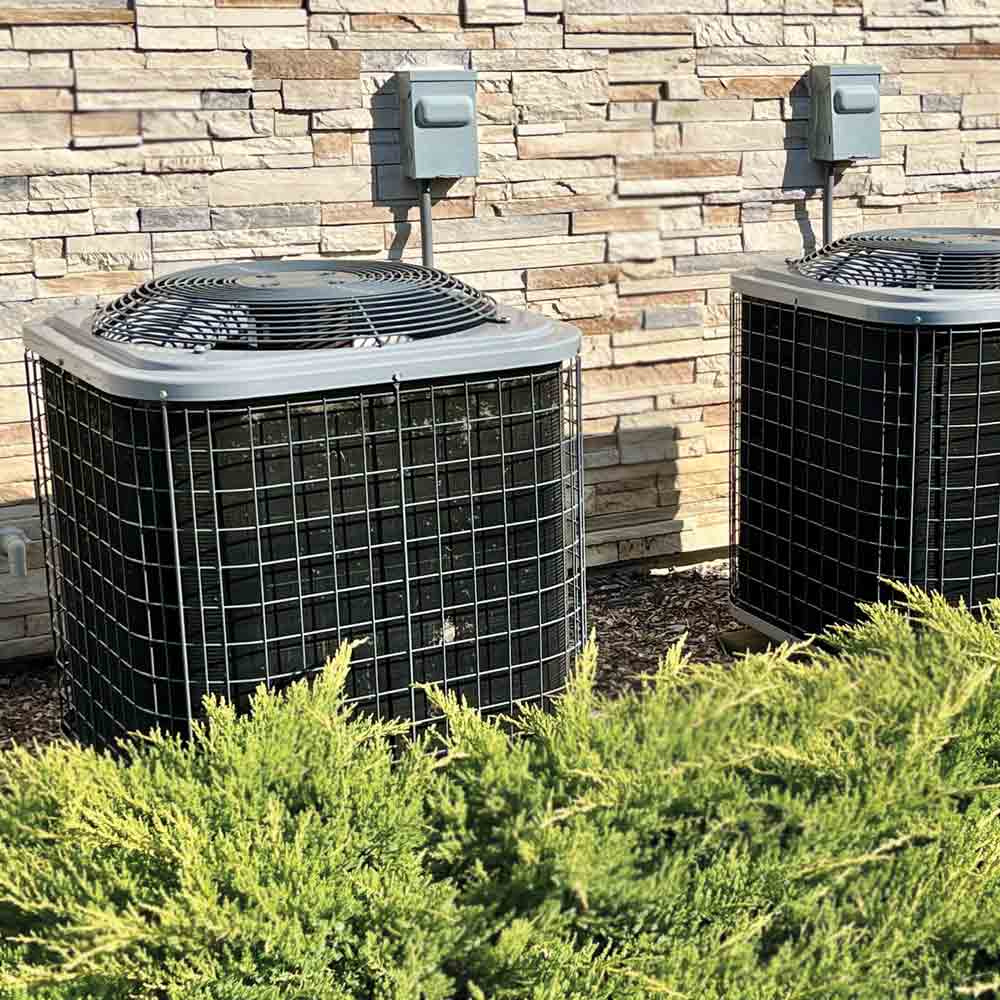 Two AC units by stone wall, shrubs in foreground.