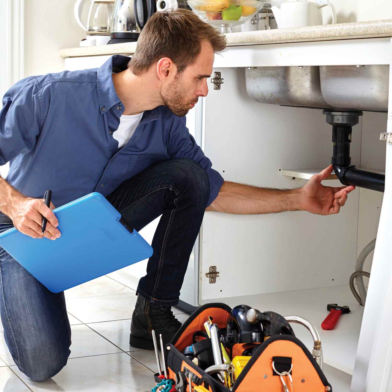 Technician checks kitchen sink plumbing with clipboard; tool bag nearby.